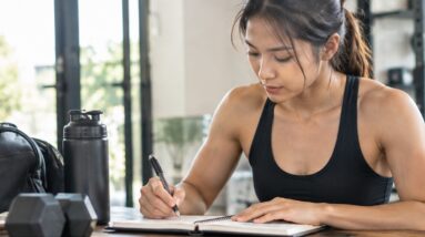 Woman writing in a fitness journal at a gym table, dumbbells and gym bag nearby, tracking her workout progress